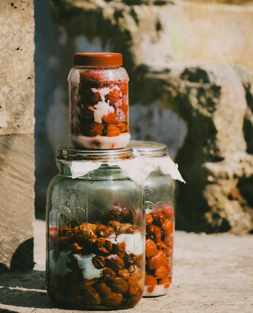 Close-up of glass jars with fermented fruits and sugar on rustic stone surface outdoors.